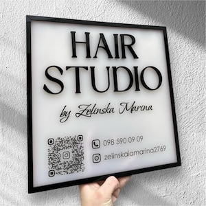 Puede incluir: Un letrero cuadrado con marco negro y fondo blanco. Las palabras "HAIR STUDIO" están en letras negras en negrita. Debajo, se lee "by Zelinska Marina". También se muestra un código QR e información de contacto.