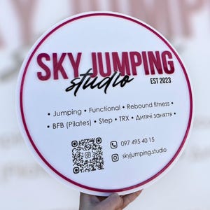 Puede incluir: Un letrero redondo blanco con borde magenta y texto. Las palabras "SKY JUMPING studio" son magenta, con "EST 2023" en la esquina superior derecha. Debajo hay viñetas con términos de fitness, un código QR e información de contacto.