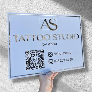 Puede incluir: Un letrero rectangular con fondo azul claro y marco plateado. El letrero dice "AS TATTOO STUDIO by Aisha" en letras plateadas. Incluye un código QR, nombre de usuario de Instagram y un número de teléfono.