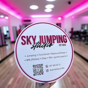 Puede incluir: Un letrero redondo blanco con borde granate y texto "SKY JUMPING studio EST 2023". El letrero también enumera tipos de clases de fitness e información de contacto, incluido un código QR y un perfil de Instagram.