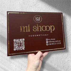 Puede incluir: Un letrero rectangular marrón con borde dorado y letras doradas. El letrero dice "mi shoop - cosmetics" e incluye un código QR e información de contacto. El letrero está sostenido por una mano.