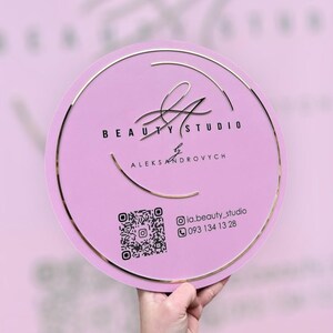 Puede incluir: Un letrero redondo de color lila claro con borde dorado y texto. El letrero dice "BEAUTY STUDIO" y "ALEKSANDROVYCH" en negro. También se incluye un código QR e información de contacto.