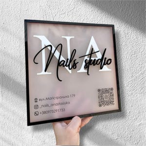 Puede incluir: Un letrero enmarcado para un estudio de uñas. El letrero presenta las letras "NA" en blanco, con "Nails studio" en negro en cursiva. También se incluye información de contacto y un código QR.