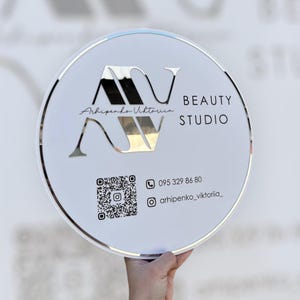 Puede incluir: Un letrero redondo blanco con un borde plateado. El letrero presenta las letras espejadas "AV" y las palabras "BEAUTY STUDIO". Debajo hay un código QR, un número de teléfono y un identificador de Instagram.