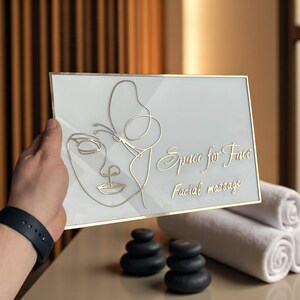 Puede incluir: Un letrero rectangular con marco dorado y fondo blanco. El letrero presenta un contorno dorado de un rostro con una mariposa y las palabras "Space for Face, Facial massage". El letrero es sostenido por una persona.