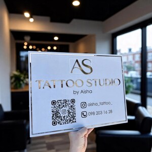 Puede incluir: Un letrero blanco con letras doradas que dice "AS TATTOO STUDIO by Aisha". El letrero incluye un código QR, un nombre de usuario de Instagram y un número de teléfono. El letrero se sostiene en una habitación bien iluminada.