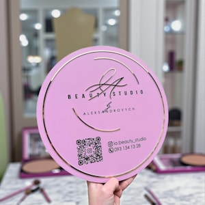 Puede incluir: Un letrero redondo rosa con borde y letras doradas. El letrero dice "BEAUTY STUDIO" y "ALEKSANDROVYCH" en letras doradas. También se incluye un código QR e información de contacto. El letrero es sostenido por una persona.