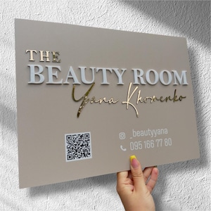 Puede incluir: Un letrero rectangular con las palabras "THE BEAUTY ROOM" en letras blancas en relieve y "Yana Khomenko" en escritura dorada. El letrero también incluye un código QR, un nombre de usuario de Instagram y un número de teléfono.