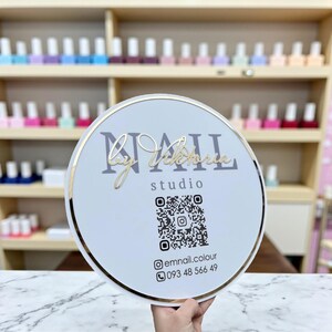 Puede incluir: Un letrero redondo blanco con borde dorado, con el texto "NAIL by Victoria studio". El letrero incluye un código QR e información de contacto. El fondo muestra estantes de botellas de esmalte de uñas.