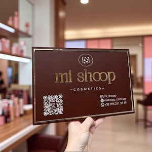 Puede incluir: Un letrero rectangular marrón con letras doradas y un borde dorado. El letrero dice "ini shoop - cosmetics-" e incluye un código QR e información de contacto. El letrero se sostiene frente a un fondo borroso.