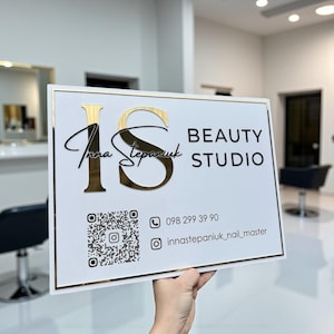 Puede incluir: Un letrero rectangular blanco con borde dorado y letras doradas que dice "IS BEAUTY STUDIO". El letrero incluye un código QR, un número de teléfono y un nombre de usuario de Instagram. El letrero se sostiene en una habitación luminosa.