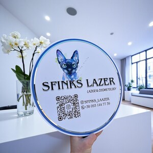 Puede incluir: Un letrero redondo con borde azul y plateado, con una ilustración de un gato azul. El letrero dice "SFINKS LAZER" con "LAZER & COSMETOLOGY" debajo. Incluye un código QR e información de redes sociales.