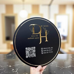 Puede incluir: Un letrero redondo negro con borde dorado y letras doradas. El letrero dice "DIANA HAIR aesthetic space" con un logotipo "DH" dorado. También se incluye un código QR e información de contacto.