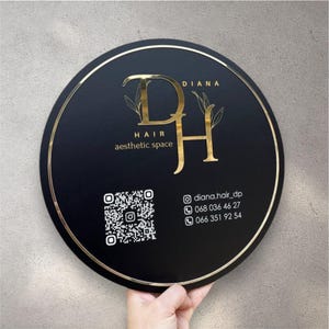 Puede incluir: Un letrero redondo negro con borde dorado y letras doradas. El letrero dice "DIANA" en la parte superior, con "DH" en letras doradas grandes en el centro. Debajo de las letras están las palabras "HAIR aesthetic space". Un código QR y la información de contacto están en la parte inferior.