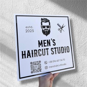 Puede incluir: Un letrero cuadrado blanco con borde negro, con el texto "MEN'S HAIRCUT STUDIO" en negrita. El letrero incluye una ilustración de barbero, tijeras y un código QR. También se muestra el año de fundación del establecimiento, 2025.