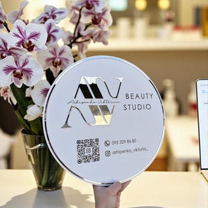 Puede incluir: Un letrero redondo blanco con borde plateado y el texto "AV BEAUTY STUDIO". El letrero también incluye un código QR e información de contacto. Un jarrón de orquídeas blancas y moradas está en el fondo.
