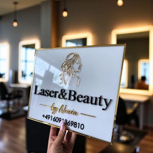 Puede incluir: Un letrero blanco rectangular con borde dorado, que muestra las palabras "Laser & Beauty" en negro. Presenta un contorno dorado del rostro de una mujer y el texto dorado "by Navin" y un número de teléfono.