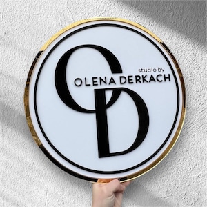 Puede incluir: Un letrero redondo blanco con un borde dorado y las letras negras "CD" en una fuente estilizada. Las palabras "studio by OLENA DERKACH" están encima de las letras. El letrero está sostenido por una mano.