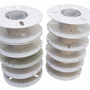 Puede incluir: Dos pilas verticales de bobinas de filamento de plástico blanco vacías. Cada bobina presenta un diseño circular con un núcleo central, múltiples perforaciones pequeñas en las superficies planas y ranuras rectangulares a lo largo de los bordes.