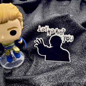 May include: A Funko Pop figure of a superhero with a blonde hairstyle, blue and gold costume, and a sticker with the text "LET ME HELP YOU" and a silhouette of a person reaching out.