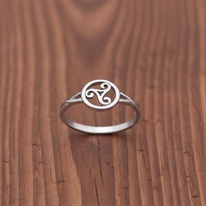 Peut inclure: Une bague en argent ornée d'un triskèle celtique. La bague présente un motif circulaire avec le symbole à trois spirales, monté sur un fin anneau. La bague est présentée sur une surface en bois.