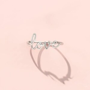 Könnte beinhalten: Ein silberner Ring mit dem Wort "love" in Schreibschrift. Der Ring liegt vor einem hellrosa Hintergrund und wirft einen Schatten. Ein zartes Schmuckstück.