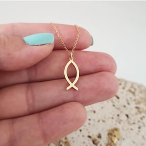 Puede incluir: Un collar dorado con un colgante con símbolo de pez. El colgante es un contorno estilizado de un pez, suspendido de una delicada cadena. El collar se sostiene en la mano de una persona con esmalte de uñas azul claro.