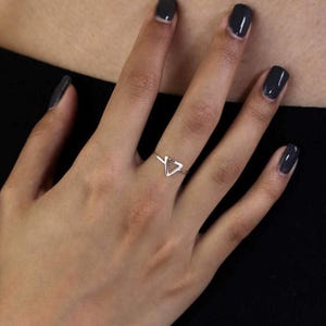 Peut inclure: Une bague fine en métal poli présentant une petite forme géométrique triangulaire ouverte centrée sur l'anneau. La bague est portée au majeur, mettant en valeur un design simple et élancé avec une finition de surface lisse et réfléchissante.