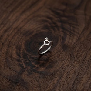 May include: A silver-toned ring featuring a circular band and a prominent male gender symbol at the top. The metal has a smooth, polished finish, and the design consists of a simple, thin band that connects to the circular icon with an upward arrow.
