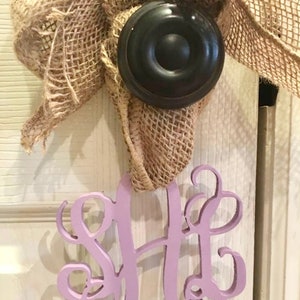Puede incluir: Un monograma de madera morado con las letras "SH" en una fuente de escritura decorativa. El monograma está colgado de un lazo de arpillera atado alrededor de un pomo de puerta negro.