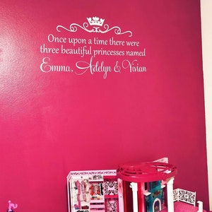 Puede incluir: Una pared rosa con letras blancas que dicen "&Eacute;rase una vez, hab&iacute;a tres hermosas princesas llamadas Emma, Adelyn y Vivian". Una casa de mu&ntilde;ecas rosa y blanca est&aacute; en primer plano.
