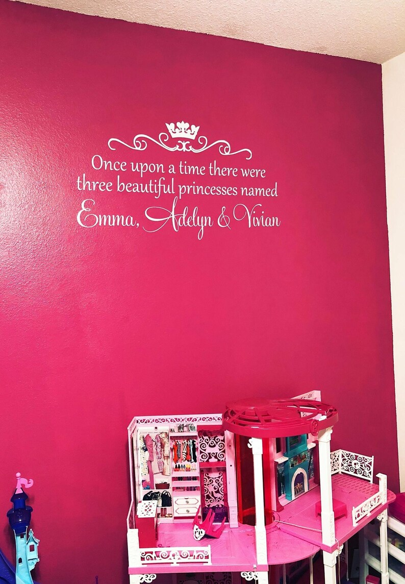 Puede incluir: Una pared rosa con letras blancas que dicen "&Eacute;rase una vez, hab&iacute;a tres hermosas princesas llamadas Emma, Adelyn y Vivian". Una casa de mu&ntilde;ecas rosa y blanca est&aacute; en primer plano.