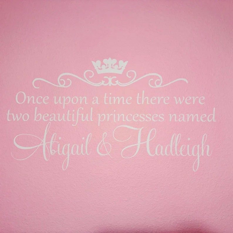 Puede incluir: Pared rosa con una calcoman&iacute;a de vinilo blanco que dice "&Eacute;rase una vez dos hermosas princesas llamadas Abigail & Hadleigh". La calcoman&iacute;a presenta una corona y un adorno decorativo.