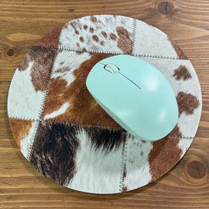 May include: A round mousepad with a patchwork design of brown, white, and tan cowhide. A light blue wireless computer mouse rests on the mousepad. The mousepad has a stitched edge.