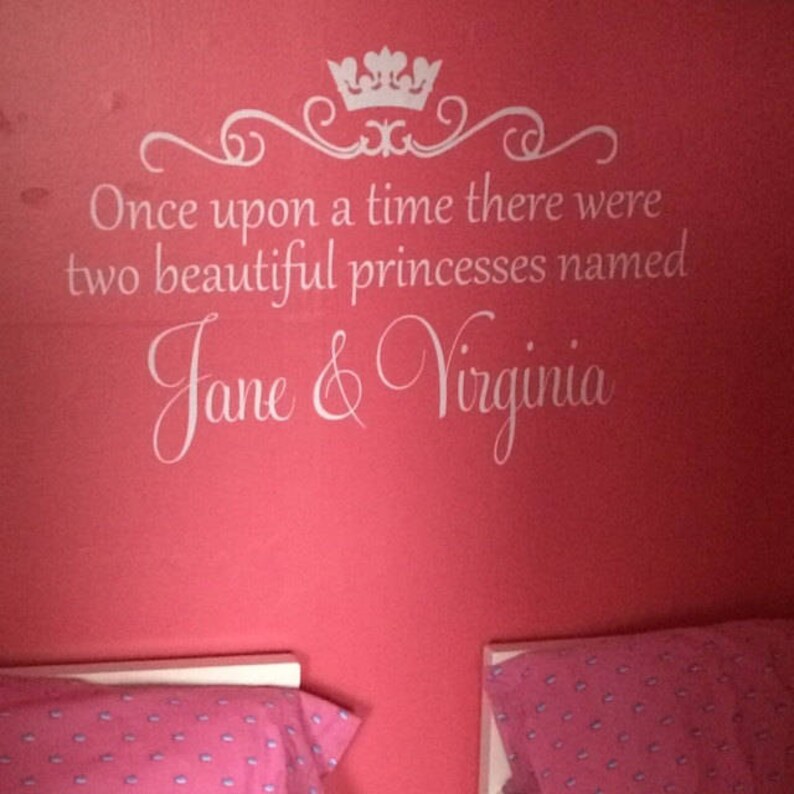 Puede incluir: Una pared rosa con una calcoman&iacute;a de vinilo blanco que dice "&Eacute;rase una vez dos hermosas princesas llamadas Jane & Virginia". La calcoman&iacute;a presenta una corona y un dise&ntilde;o de desplazamiento decorativo. Dos camas individuales con ropa de cama estampada en rosa y blanco est&aacute;n en primer plano.
