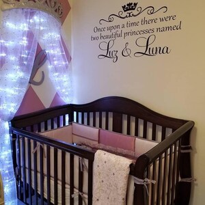 Puede incluir: Una cuna de madera marr&oacute;n con un juego de ropa de cama blanco y rosa. La cuna est&aacute; en una habitaci&oacute;n de beb&eacute; con una pared de patr&oacute;n de diamantes rosa y blanco. Un dosel blanco con luces est&aacute; colgado de la pared sobre la cuna. Una calcoman&iacute;a de pared con el texto "&Eacute;rase una vez dos hermosas princesas llamadas Luz & Luna" est&aacute; en la pared.