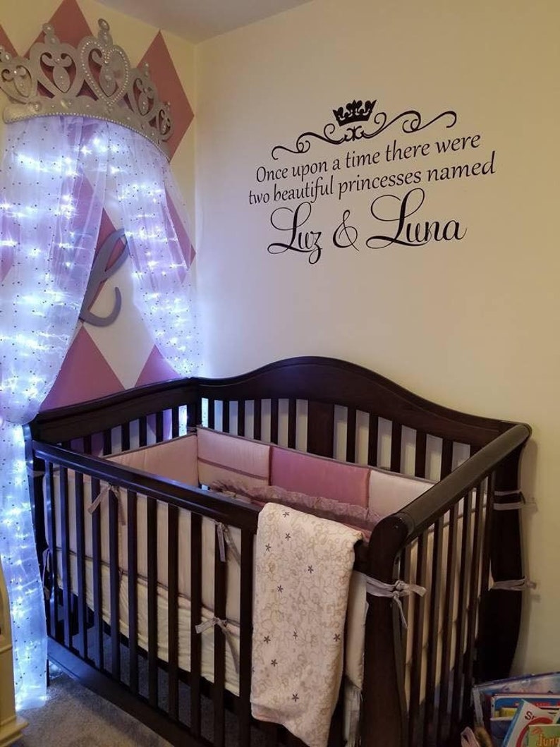Puede incluir: Una cuna de madera marr&oacute;n con un juego de ropa de cama blanco y rosa. La cuna est&aacute; en una habitaci&oacute;n de beb&eacute; con una pared de patr&oacute;n de diamantes rosa y blanco. Un dosel blanco con luces est&aacute; colgado de la pared sobre la cuna. Una calcoman&iacute;a de pared con el texto "&Eacute;rase una vez dos hermosas princesas llamadas Luz & Luna" est&aacute; en la pared.