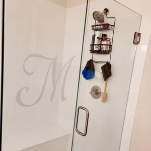 Può includere: Un portasapone da doccia in metallo nero appeso al soffione, con due ripiani in filo metallico per vari flaconi, ganci per accessori e una porta in vetro con una grande lettera M incisa sulla parete interna rivestita di piastrelle bianche.