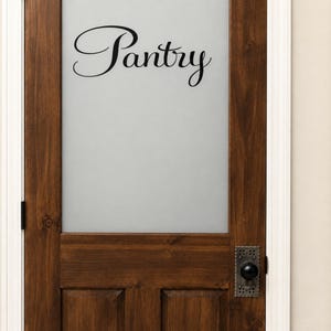 May include: A dark brown wooden door with a large rectangular frosted glass panel in the center. The word Pantry is written in black cursive script on the glass. A rectangular metal backplate with a round black knob is attached to the door.