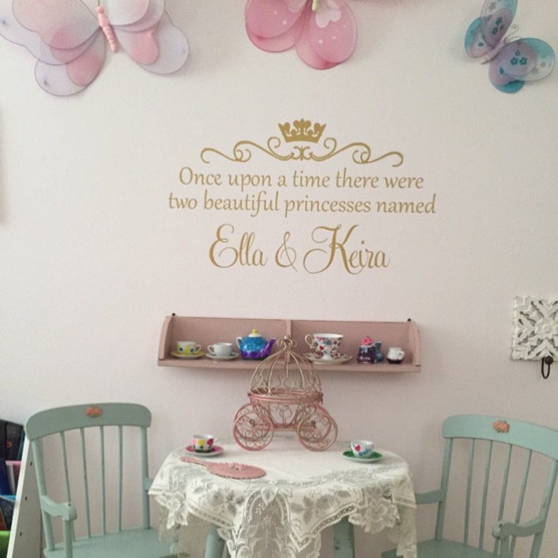 Puede incluir: Una calcoman&iacute;a de pared de brillo dorado con el texto "Once upon a time there were two beautiful princesses named Ella & Keira" con una corona sobre el texto. La calcoman&iacute;a est&aacute; en una pared blanca con mariposas rosas y un estante blanco con tazas de t&eacute; y una tetera.
