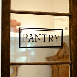 May include: A black vinyl decal applied to a glass door panel featuring the word PANTRY in a serif font, enclosed within a double-lined rectangular border. The decal is centered on the transparent glass pane of a dark brown wooden door frame.