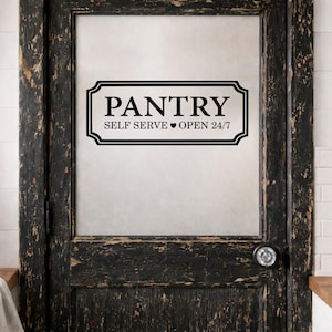 May include: A black and white pantry sign is fixed to a distressed, dark wood door. The sign reads "PANTRY" above "SELF SERVE OPEN 24/7" in a decorative frame. The door has a glass panel and a round doorknob.