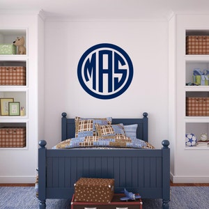 Op de afbeelding: Een marineblauwe monogram sticker met de letters "MAS" in een cirkel op een witte muur in een slaapkamer. De slaapkamer heeft een blauw bed met een geruit beddengoed en een bruin-witte geblokte opbergdoos.