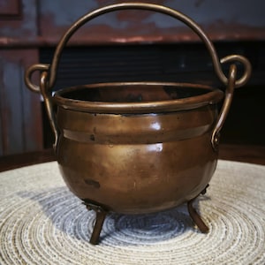 Può includere: Un calderone in rame vintage con manico e tre piccole gambe. Il calderone ha un corpo arrotondato e una patina marrone scuro. Il manico è arcuato e attaccato ai lati della pentola. Il calderone è appoggiato su un sottobicchiere rotondo e tessuto.
