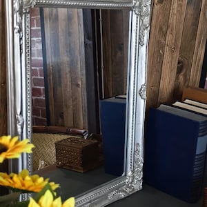 Può includere: Specchio rettangolare argentato con cornice ornata. Lo specchio riflette una stanza con pannelli di legno, una piccola scatola di legno e libri blu. Girasoli gialli in primo piano.