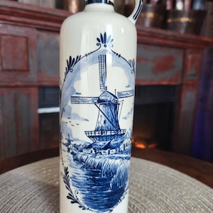 May include: A tall, white ceramic bottle with a blue windmill design. The bottle has a handle and a stopper. The windmill scene is detailed with blue accents. The bottle is approximately 12 inches tall.