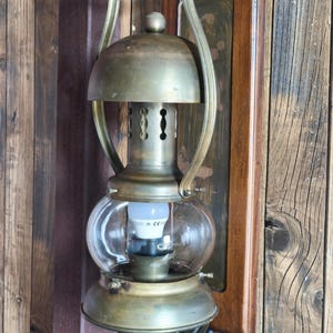 Peut inclure: Ancienne lanterne murale en laiton avec un globe en verre transparent et une poignée en bois. La lanterne est fixée sur une plaque de bois et présente un design décoratif. Le texte en bas se lit : "Not trade - a ritual of creation. Every piece, a fragment of another world."