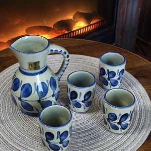 Könnte beinhalten: Ein Keramikkrug und vier passende Tassen. Der Krug hat einen Henkel und einen Ausguss. Alle Teile sind cremefarben mit blauen Blumenmustern. Das Set steht auf einem runden, gewebten Platzset.