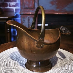 May include: A copper-colored metal coal scuttle with a curved handle and a small wooden handle on the side. The scuttle sits on a round, woven placemat. The image has a vintage aesthetic.