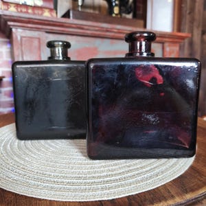 Può includere: Due bottiglie quadrate di vetro scuro con tappi arrotondati. Le bottiglie hanno un'estetica vintage, con un colore profondo, quasi nero. Le bottiglie sono esposte su un tappetino rotondo e intrecciato.